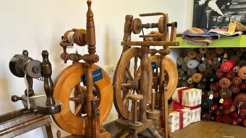Spinning wheels stand next to a stack of yarn supplies, all in different colours. Boxes are decorated with red ribbons for Christmas.