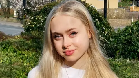 Owen Ward/BBC Head and shoulders image of Erin LeCount, who has long blonde hair and is wearing a white top, standing in front of foliage