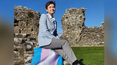 John Dunkley-Williams Gwion Williams smiling at the camera in the ruins of a castle with the blue, pink and white trans flag laid out next to him