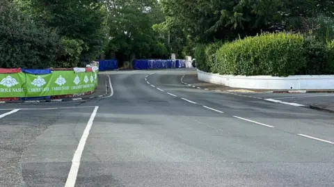 Green crash barriers on the left at Cruickshanks with more blue barriers ahead on the left and a white wall next to a pavement on the right.