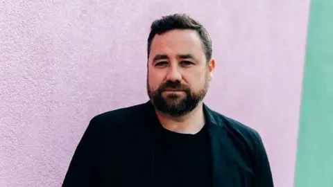 Rosie Powell A man with dark brown hair and a beard. He is wearing a black t-shirt and standing in front of a colourful wall. 