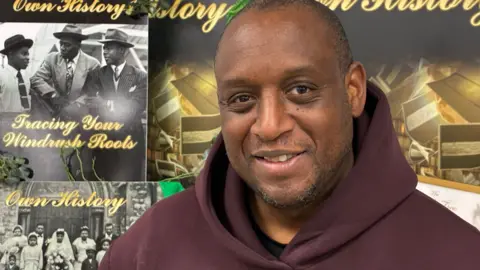A picture of Adrian Stone in front of his 'Tracing Your Windrush Roots' display. He is smiling and has a dark brown hoodie on