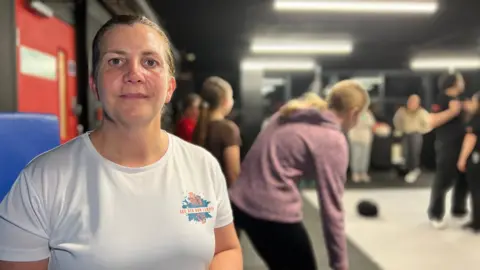 Ceri looks at the camera as the instructor shows the girls a technique behind her. She is wearing a white training top with the See Her Run logo on it. She has brown eyes and short brown hair which is tied into a pony tail.