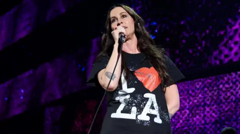 Getty Images A woman with brown hair smiling at the camera wearing black top. She is holding a microphone with the stage design behind her suggesting she is in a live performance.