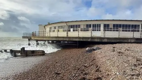 Worthing Borough Council A single storey cream building built on a beach. The image shows the building from the side, with the end of the building hanging over the sea