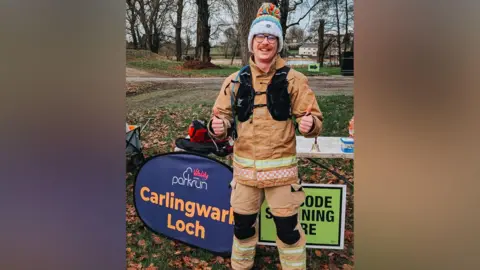 Taz Maguire A man dress in a beige, black and yellow firefighter suit is smiling at the camera with his thumbs up. There is greenery and trees behind him along with a sign that reads, "Vitality park run, Carlingwark Loch".