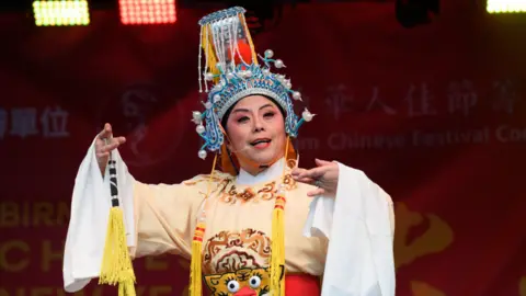 Birmingham Hippodrome A performer in a traditional Chinese opera costume stands on a stage with bright lighting overhead. The outfit includes a cream robe decorated with patterns. The backdrop is red with festival branding faintly visible behind the performer.
