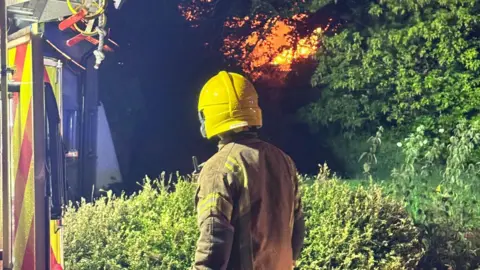 A firefighter at the scene of a fire. Orange sky with flames can be seen in the distance