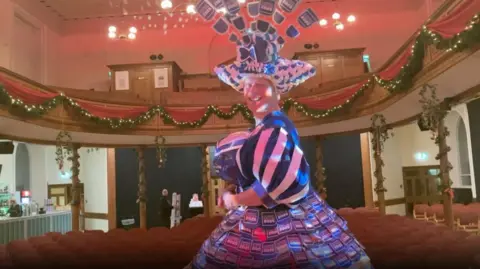 BBC Matt wearing the manifesto dress whilst standing on a stage. The dress is blue and white with a matching headpiece which features cut outs from the manifesto book. In the background, there are empty seats. 