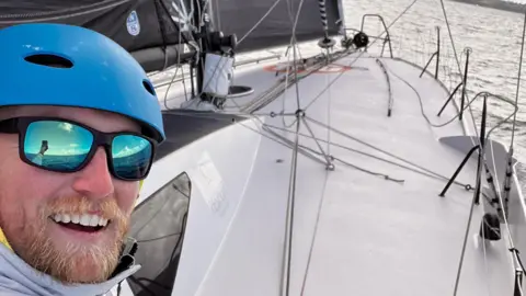A picture of a man smiling at the camera while taking a selfie. He is wearing glasses and a helmet. The boat can be seen in the background. 