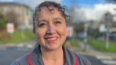 Head and shoulders of a smiling woman with grey curly hair tied back in a grey with red piping nurses uniform with a hospital in the background