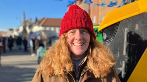 BBC/Mark Ansell Sophie Bramley is smiling looking straight ahead. She has long hair and is wearing a red hat with a brown jacket. She is stood at the side of a yellow tractor. 