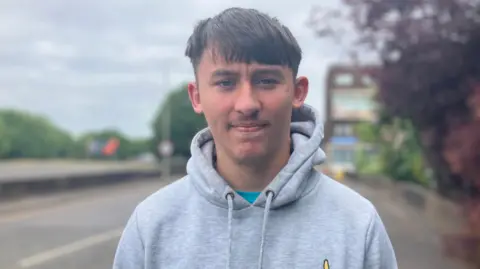 Jermaine wears a grey hooded top and stands by the side of the road next to the works.