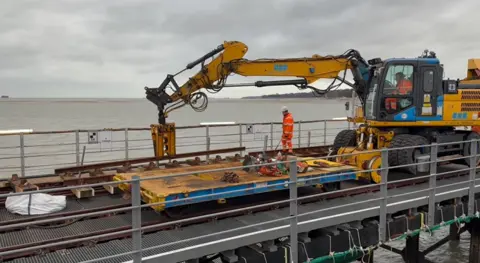 Network Rail A yellow digger holding a steel girder on the railway tracks with two engineers wearing orange work overalls.