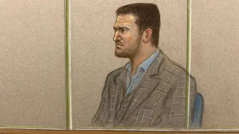 Julia Quenzler A court sketch of Tommy Robinson at Woolwich Crown Court in October 2024. He is grimacing and frowning as he looks ahead, seated at an angle. He is behind a window with two bars and is wearing a blue open-collar shirt and a brown check jacket. He has short brown hair and a neat beard and moustache.