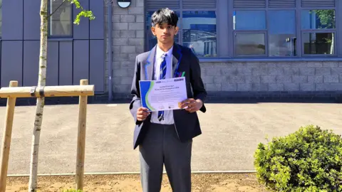 Bedford Giving Sempiyan, a pupil, wearing a blue blazer, blue trousers, a white shirt, and a tie. He is standing by a school building, holding up a certificate. He is looking straight at the camera. 
