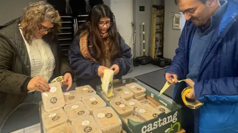 Two women, wearing dark brown coats - one younger with dark hair and glasses and the other older with blonde hair - sort out meals in plastic tubs into boxes