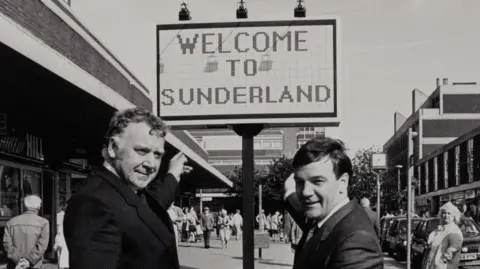 A black and white image of two men in suits pointing to a sign that reads: "Welcome to Sunderland". They are standing on a high-street bordered by shops in concrete and brick buildings. The men are smiling at the camera.