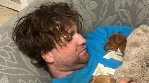 Nikita Goddard James Nunan has long curly brown hair and is lying on a grey sofa with two brown puppies on his chest. He is wearing a blue jumper with white lettering.