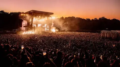 A teeming crowd of thousands holding up smartphones towards a stage lit up with yellow lights and covered with dry ice. A line of trees can be seen in darkness as the sun sets.