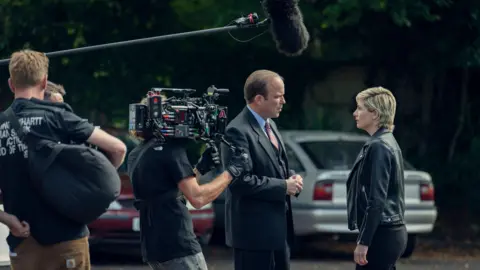 Netflix Rory Kinnear is speaking with Jodie Whittaker with a boom microphone above them and a man holding a camera stood nearby