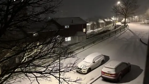 Snowy street with houses on one side and two cars, covered in snow and parked