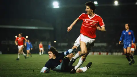 Getty Images Ian Rush, in a red Wales shirt and white shorts and socks, rounds a prone goalkeeper to score a goal in an international match, as players from both sides watch in the background