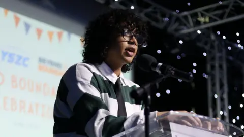 ISCRE Efaz stands at a lectern speaking into a microphone. He is standing on a stage and is wearing a green and white striped top. 