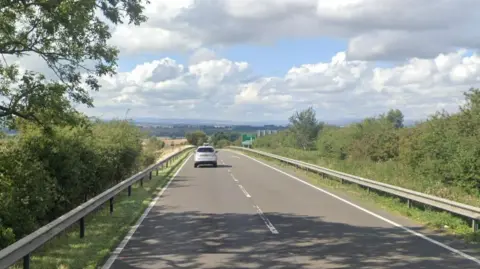 A dual carriageway road with trees on either side and a view of hills in the distance. A silver car on the road. Blue sky with clouds.