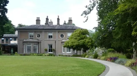 Compton Care The hospice is in a picturesque beige building with a sweeping drive and grass and garden either side of it.