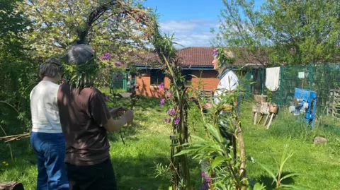 Cath Darling is in the background standing on a lawn in a garden with a brick built single-storey behind her. In the foreground of the picture, two women are standing in front of an arch made of willows and flowers.