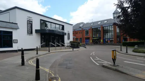 The road with double yellow lines leading to High Cross Church. Camberley Theatre on the left side in Camberley, England, UK May 28, 2022. Blue skies and clouds in the background