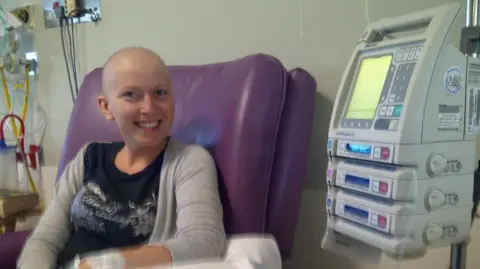 Selina Moss-Davies A woman sat in a chair undergoing cancer treatment. She is smiling at the camera. There is hospital machinery to her left, our right