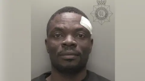 A police custody photo of Timothy Kusemi. He has short black hair and short black facial hair. He is wearing a black t-shirt and has a white plaster on his left side of his head. The wall behind him is grey.