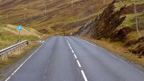 A road in Shetland with a parking sign to the left. There is banked grassland at both sides and another road leading off to the left after the parking sign