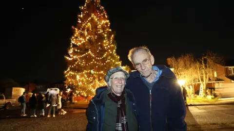 SWNS A elderly couple pose for a photo stood next to each other with a large Christmas tree behind them illuminated in warm lighting. A crowd of people are gathered behind them looking at the tree. 