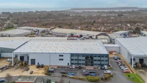 JLL An aerial view of a large grey industrial unit on a business park with houses in the background and the sea in the distance.