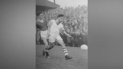 Workington AFC A black and white image of two players chasing the ball during the Workington Reds v Manchester United match at Borough Park in 1958. There is a large crowd seen close to the touchline, rising up the bank behind.