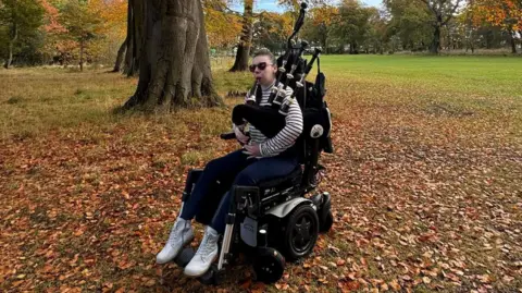Katie Robertson A woman in a wheelchair in the middle of a park playing the bagpipes on an autumn day with leaves surrounding her. 