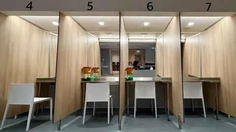 Numbered booths with a table and chair each are in a row. There is a waste bin in the foreground.