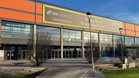 The exterior of the SSE Arena in Belfast. It is a large orange brick building with dark framed large windows and a sign above the glass doors which reads 'The SSE Arena'. There is also a large sign covering the top which says 'Ready for a rewarding night?'.