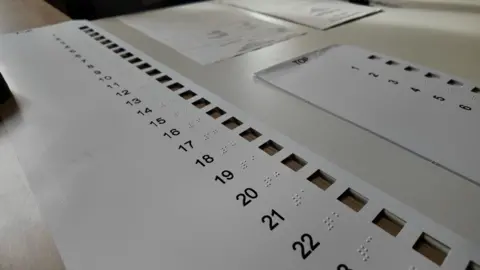 A longer overlay for the bigger ballot paper has more than 20 numbers, top to bottom with Braille beside the numbers to help the voter. The overlay is white and the numbers are in black.