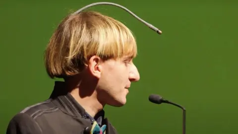 First Born Films Neil Harbisson vista lateral dele e suas antenas, que começam na parte de trás de sua cabeça e formam um arco acima de sua testa