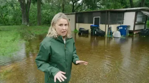 Sarah Smith delivering a report in front of a flooded house