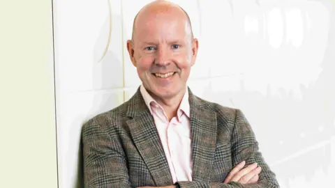 Mark Preece stands leaning against a wall with his arms folded, he is smiling and wearing a tweed jacket with a pink shirt. 