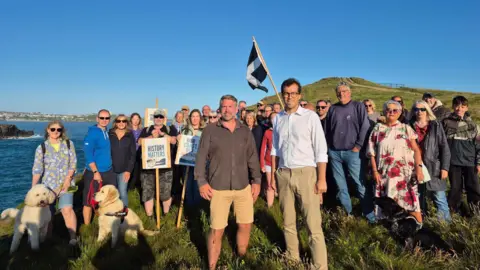 Matt Kennedy-Smith Group of supporters gathered on a coastal headland with campaign placards and a Cornish flag, overlooking the sea.
