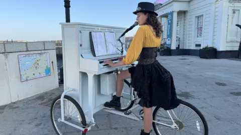 Jo Burn/BBC Chloe Marie Aston in a yellow cardigan and black dress stands sits on her bike while playing the piano.