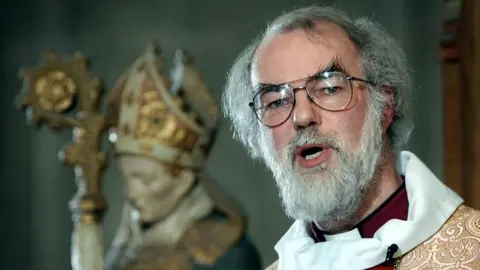 PA Media Dr Rowan WillIams gives a sermon. He has thick grey hair receding on top, and a beard and is wearing a glasses. He wears white, red and gold robes. Behind him is an ornate statue of a clerical figure