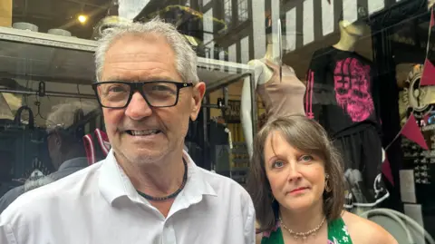 Alice Cunningham/BBC Stuart Mill and Rebecca Eaks smile at the camera in front of their shop. Stuart has short grey hair and wears a white shirt. Rebecca has short brown hair and a green top.