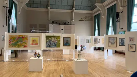 An art exhibition displayed in a large hall, with paintings and sculptures arranged on freestanding white panels and plinths.